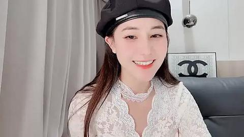 Media: Video of a young Asian woman with fair skin, long dark hair, wearing a black beret, white lace blouse, and smiling. Background features a grey curtain and a \"CHANEL\" sign.