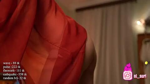 Media: Video of a woman's upper body in a red, sheer blouse, revealing her light skin and small breasts. Background shows a dimly lit room with beige curtains and a wooden door.