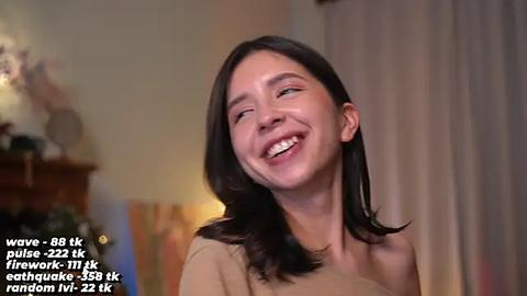 Media: Video of a smiling Asian woman with straight, shoulder-length black hair, wearing a beige top, in a dimly lit room with a wooden dresser and a curtain.