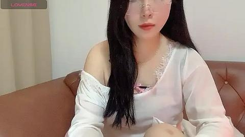 Media: Video of an East Asian woman with long black hair and fair skin, wearing glasses and a white off-shoulder top, seated on a brown leather couch in a minimalist room.