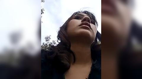 Media: A close-up video of a young woman with dark, wavy hair and fair skin, looking up with a serene expression. Background is out of focus, with hints of trees and sky.