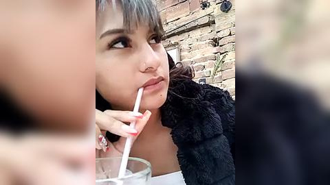 Media: Video of a young woman with straight black hair, light skin, and a white straw in her mouth, sitting outdoors next to a brick wall. She wears a dark, textured jacket.