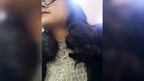 Media: Video of a young woman with long, wavy black hair, wearing black-framed glasses and a gray, patterned turtleneck sweater, standing in a dimly lit room.