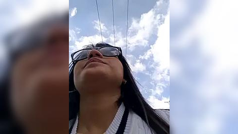 Media: A low-angle video of a young Asian woman with long black hair and glasses, wearing a white cardigan, looking up at the sky filled with clouds.