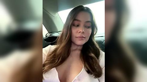 Media: A blurry video shows a young woman with long brown hair, wearing a low-cut white top, sitting in a car with the window down, looking out.