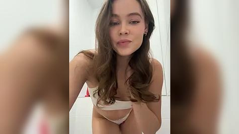 Media: Video of a young Caucasian woman with long brown hair, wearing a white bra and matching panties, leaning forward in a bathroom.