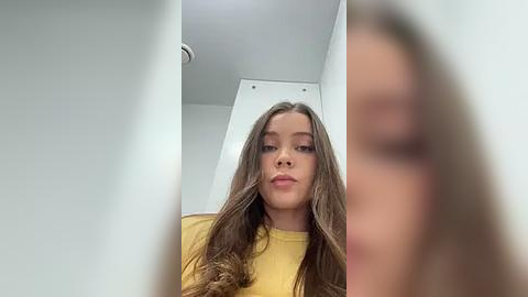 Media: Video of a young woman with long brown hair and fair skin, wearing a yellow shirt, looking directly into the camera from a low angle, with blurred background.