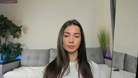 Media: Video of a young woman with long, straight brown hair, fair skin, and a slender physique, wearing a white top. She's seated on a modern gray sectional sofa with decorative pillows in a minimalist living room.