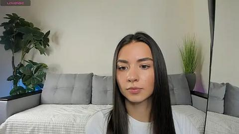 Media: Video of a young, light-skinned Latina woman with long, straight black hair, wearing a white T-shirt, sitting on a gray sectional sofa. The background features a green potted plant and a tall, thin decorative plant.