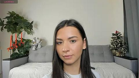 Media: A video of a young woman with long dark hair, fair skin, and light makeup, sitting on a grey couch in a modern living room with green plants and decorative candles.