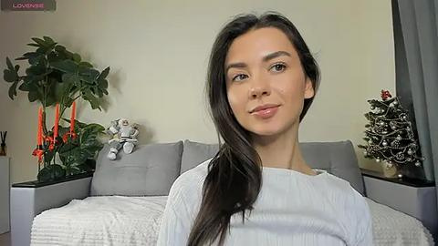 Media: Video of a young woman with long dark hair, wearing a white sweater, standing in a modern, cozy living room with a gray couch, potted plants, and Christmas decorations.