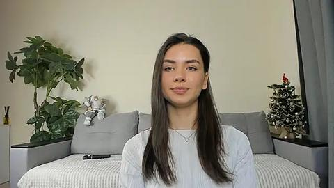 Media: Video of a young woman with straight, dark hair, wearing a white sweater, standing in a modern living room with a gray couch, green plants, a Christmas tree, and a teddy bear.