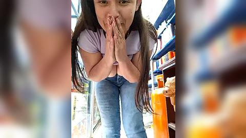 Media: A video of a young Asian woman with long black hair, wearing a light purple shirt and blue jeans, hands clasped in prayer, standing in a brightly lit convenience store with colorful snacks and beverages in the background.