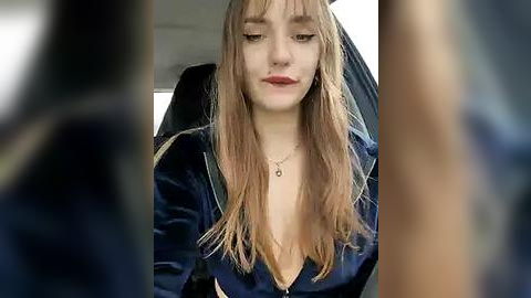 Media: Video of a young woman with long, straight, light brown hair, wearing a dark blue velvet jacket, sitting in a car, with blurred background.