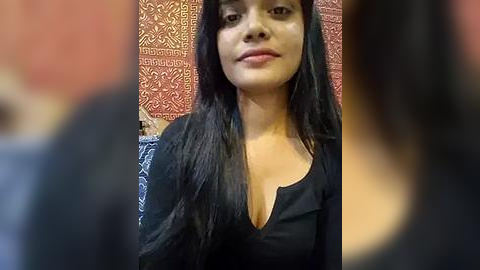 Media: Video of a South Asian woman with long black hair, wearing a black top, smiling, against a red-patterned wall. Background slightly blurred.