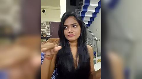 Media: Video of a young South Asian woman with long black hair, wearing a black sleeveless top, gesturing with her hand. Background includes modern staircase and wall art.