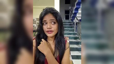 Media: Video of a young Indian woman with long black hair, wearing a sleeveless red dress, sitting on a staircase with a worried expression.