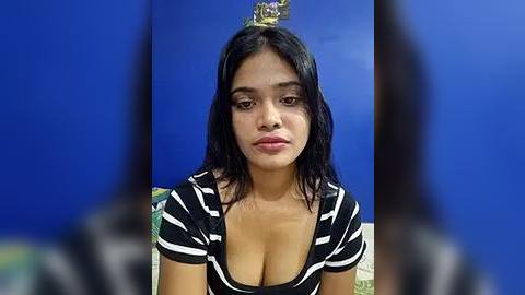 Media: Video of a young woman with long black hair, wearing a black and white striped top, sitting against a blue wall. Her expression is neutral, and she appears to be in a casual indoor setting.