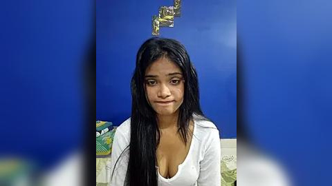 Media: Video of a young woman with long black hair, fair skin, wearing a low-cut white top, against a blue wall with a golden, abstract art piece above her head.