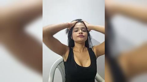 Media: A video of a Latina woman with medium skin tone, long black hair, and medium breasts, wearing a black tank top, sitting with hands on her head in a white room.