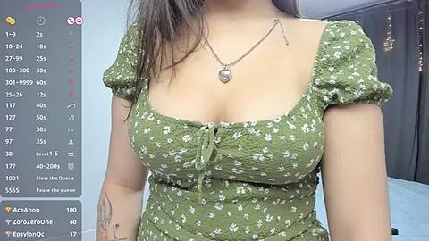 Media: A video of a young woman with light brown skin and long dark hair, wearing a green floral dress with puffed sleeves, and a heart pendant necklace. Background shows a dimly lit room with a black curtain.