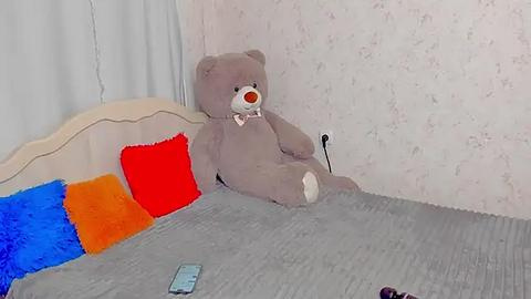 Media: Video of a beige teddy bear with a pink nose and white paws, sitting on a gray bed with colorful pillows (red, blue, and orange) against a white floral wallpaper background.