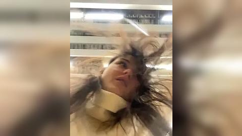 Media: Video of a woman with long, dark hair, wearing a white sweater, with her hair flying around her head in a chaotic manner, taken indoors with a blurred background.