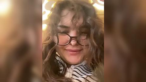 Media: A video of a young woman with curly, shoulder-length hair, wearing black-rimmed glasses, smiling. She has a light complexion and is dressed in a striped blouse. The background is blurred, with warm lighting.