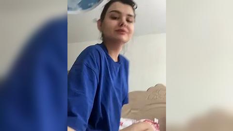 Media: Video of a young woman with light skin, brown hair, wearing a blue shirt, standing in a bedroom with beige walls and a bed with pink sheets.