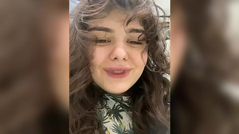 Media: A close-up video of a young woman with curly, voluminous dark brown hair, fair skin, and a slight smile. She wears a floral-patterned top. The background is blurred, focusing attention on her expressive face.