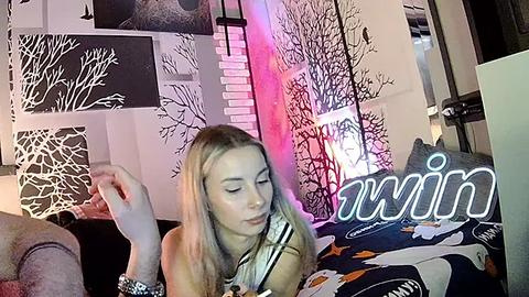Media: Video of a blonde woman in a striped top, lying on a bed with a colorful duvet, surrounded by black and white abstract art and a glowing \"avin\" sign.