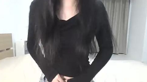 Media: Video of a person with long black hair, wearing a black sweater, standing in a bedroom with beige curtains and a light-colored bed.