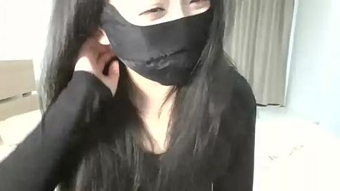 Media: Video of an East Asian woman with long black hair, wearing a black face mask and long-sleeved black top, adjusting her mask in a modern, minimalist room with white walls and a beige curtain.