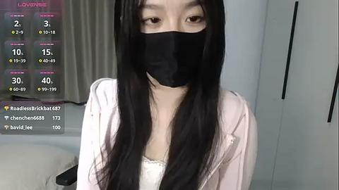 Media: Video of a young Asian woman with long black hair, wearing a black face mask, white hoodie, and white tank top, standing indoors, displaying a live stream overlay with viewer count and chat messages.