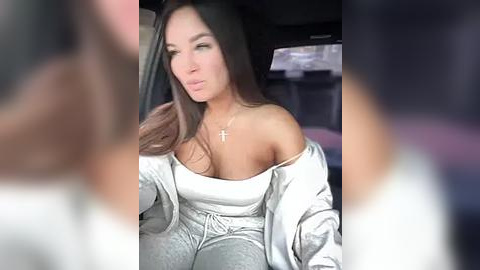 Media: Video of a woman with long dark hair, wearing an off-shoulder white top and silver jacket, seated in a car.