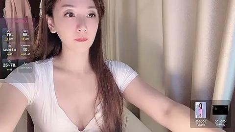 Media: Video of an Asian woman with long brown hair, fair skin, and a slim physique, wearing a white, low-cut V-neck top. She's indoors, possibly in a bathroom, with beige curtains in the background.