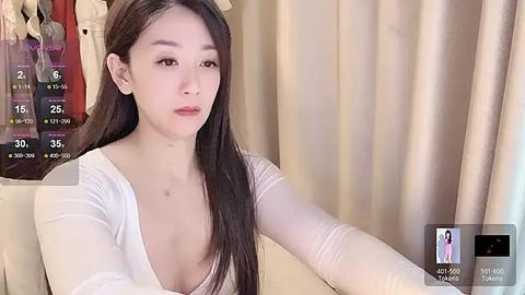 Media: Video of an East Asian woman with long black hair, fair skin, and small breasts, wearing a white long-sleeve top, sitting against beige curtains.