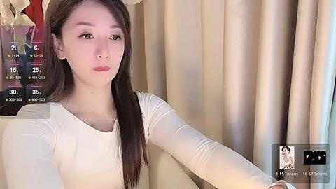 Media: Video of a young Asian woman with straight, dark brown hair, light skin, and a slim physique, wearing a white long-sleeve top. She is indoors, seated against a beige curtain background, looking contemplative.