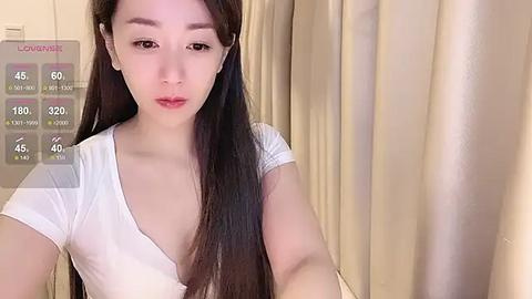 Media: A video of an Asian woman with long, straight black hair, fair skin, and a slender physique, wearing a white, low-cut top. She stands against beige curtains.