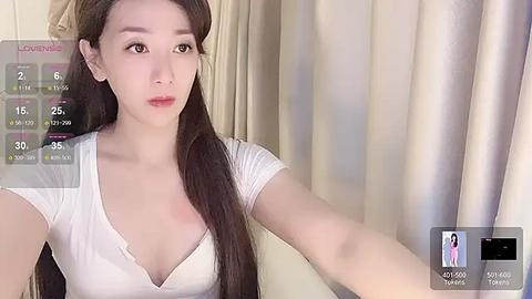 Media: A video of an East Asian woman with fair skin, long dark hair, and a white V-neck top. She has a neutral expression and stands in front of beige curtains.