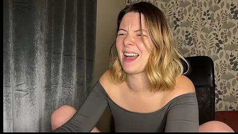 Media: Video of a laughing woman with shoulder-length, blonde ombre hair, wearing a grey off-shoulder top, seated in a room with grey curtains and floral-patterned wallpaper.