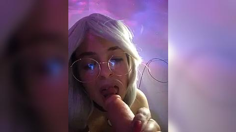 Media: Video of a light-skinned woman with long platinum blonde hair and glasses, performing oral sex on a large, circumcised penis in a dimly lit room.