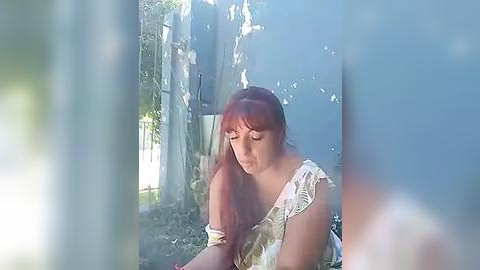 Media: Video of a young woman with long, straight red hair, wearing a white, off-shoulder floral dress, sitting in a sunlit, natural forest setting with tall trees and dappled sunlight filtering through the leaves.