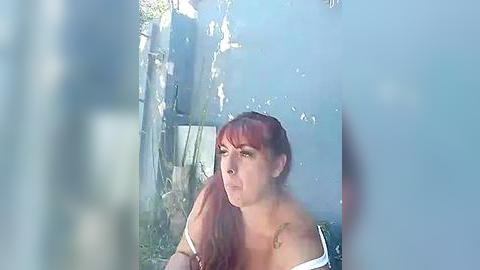 Media: Video of a fair-skinned, red-haired woman with bangs, wearing a white off-shoulder top, looking contemplative through a glass door. Background features a blurred outdoor setting with sunlight filtering through.