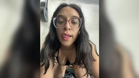 Media: Video of a young, slender Latina woman with long, dark hair and glasses, wearing a black bra, licking her lips seductively.