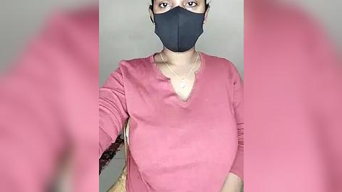 Media: Video of a woman with medium skin tone, wearing a black face mask, pink long-sleeved top, and braided hair, standing indoors with blurred background.