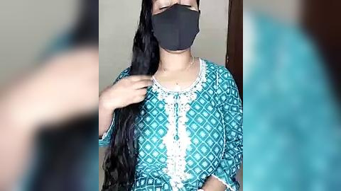 Media: Video of a woman with long black hair, wearing a teal and white patterned kurta, black mask, and a teal saree, with a blurred background.