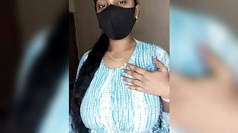 Media: Video of a young woman with medium-dark skin, wearing a blue and white striped dress, black face mask, and a black headband. She has a slender build and large breasts. The background is blurred.