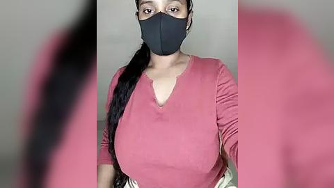 Media: Video of a woman with medium skin tone, wearing a black face mask, long black hair in a ponytail, and a pink long-sleeve top. The background is blurred, with a pink filter applied.