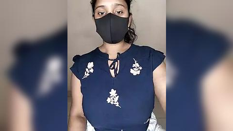 Media: Video of a woman with medium skin tone, wearing a black face mask, navy-blue blouse with white floral patterns, and a black bow tie.